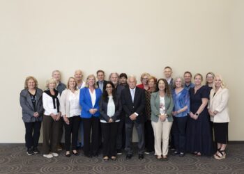 A group of about 18 professionally dressed adults standing in a three-row formation against a large, plain, off-white wall. The group consists of men and women of various ages, smiling and looking toward the camera. Many are wearing business casual attire, including blazers, cardigans, and blouses in shades of blue, beige, grey, and black. They are standing on a dark grey carpet with a large, interlocking circular pattern.