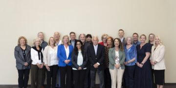A group of about 18 professionally dressed adults standing in a three-row formation against a large, plain, off-white wall. The group consists of men and women of various ages, smiling and looking toward the camera. Many are wearing business casual attire, including blazers, cardigans, and blouses in shades of blue, beige, grey, and black. They are standing on a dark grey carpet with a large, interlocking circular pattern.