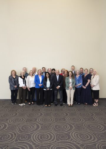 A group of about 18 professionally dressed adults standing in a three-row formation against a large, plain, off-white wall. The group consists of men and women of various ages, smiling and looking toward the camera. Many are wearing business casual attire, including blazers, cardigans, and blouses in shades of blue, beige, grey, and black. They are standing on a dark grey carpet with a large, interlocking circular pattern.