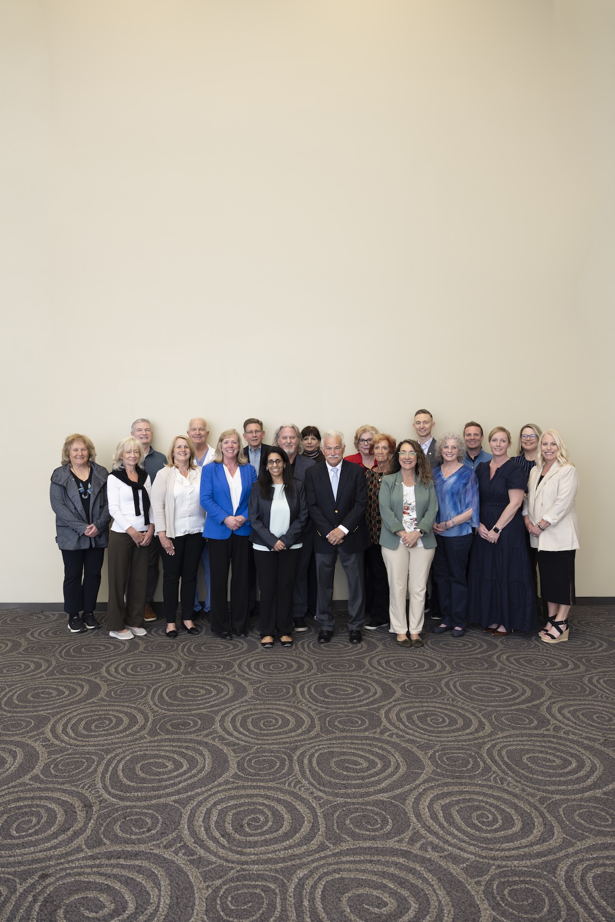 A group of about 18 professionally dressed adults standing in a three-row formation against a large, plain, off-white wall. The group consists of men and women of various ages, smiling and looking toward the camera. Many are wearing business casual attire, including blazers, cardigans, and blouses in shades of blue, beige, grey, and black. They are standing on a dark grey carpet with a large, interlocking circular pattern.