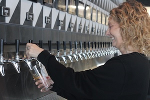 A side-profile, medium shot of a smiling woman with curly blonde hair using a self-pour beverage wall at Westfield Collective. She is wearing a black long-sleeved shirt and is holding a clear glass under one of the many chrome taps mounted on a stainless steel backsplash. Above the taps, a wall features a modern, black-and-white geometric triangular pattern and a row of digital screens. The lighting is bright and clear, capturing the interactive and modern atmosphere of the space in Westfield, Indiana.