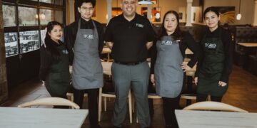 A professional group portrait of five employees at a Toro Mexican restaurant, standing between tables in a clean, contemporary setting.