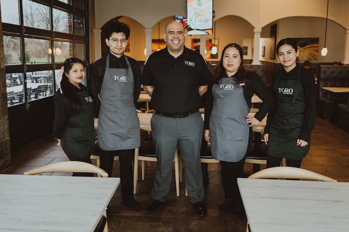A professional group portrait of five employees at a Toro Mexican restaurant, standing between tables in a clean, contemporary setting.