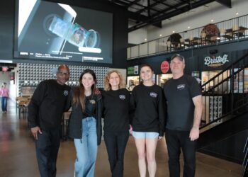 A medium full-shot of a diverse group of five smiling people standing in the center of the modern Westfield Collective food hall. Three men and two women are gathered together, with several wearing black t-shirts featuring the "Westfield Collective" logo. The industrial-style space has polished concrete floors, high black ceilings with exposed white ductwork, and a second-level mezzanine with a metal railing. In the background, a massive 25-foot LED screen displays sports content, and signs for local food vendors like Bristol's Burgers and Fork & Fire are visible on the right.