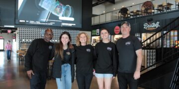 A medium full-shot of a diverse group of five smiling people standing in the center of the modern Westfield Collective food hall. Three men and two women are gathered together, with several wearing black t-shirts featuring the "Westfield Collective" logo. The industrial-style space has polished concrete floors, high black ceilings with exposed white ductwork, and a second-level mezzanine with a metal railing. In the background, a massive 25-foot LED screen displays sports content, and signs for local food vendors like Bristol's Burgers and Fork & Fire are visible on the right.