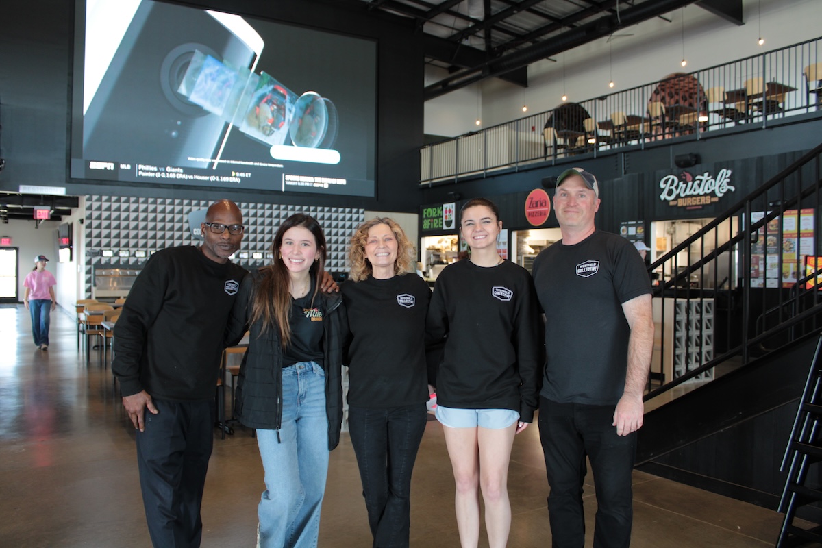 A medium full-shot of a diverse group of five smiling people standing in the center of the modern Westfield Collective food hall. Three men and two women are gathered together, with several wearing black t-shirts featuring the "Westfield Collective" logo. The industrial-style space has polished concrete floors, high black ceilings with exposed white ductwork, and a second-level mezzanine with a metal railing. In the background, a massive 25-foot LED screen displays sports content, and signs for local food vendors like Bristol's Burgers and Fork & Fire are visible on the right.