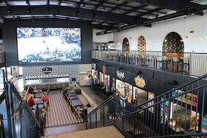 A high-angle, wide-perspective shot looking down into the main hall of the Westfield Collective in Westfield, Indiana. The industrial-style space features high black ceilings with exposed ductwork and a second-level mezzanine with a black metal railing. In the center is a 25-foot LED screen displaying a hockey game. Below the screen is a long, modern bar and several dining tables. The right side of the main floor features vendor stalls for Bristol's Burgers and Fork & Fire, and a black metal staircase leads to the upper level. The space is brightly lit by recessed square ceiling lights, highlighting the polished concrete floors.