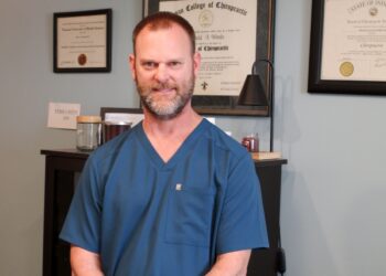 A professional portrait of Dr. Ronald Woods, a chiropractor with a short beard, wearing navy blue FIGS scrubs. He is standing in his office with his hands clasped in front of him. In the background, his diploma from Logan College of Chiropractic and his license from the State of Indiana are displayed on a light blue wall.