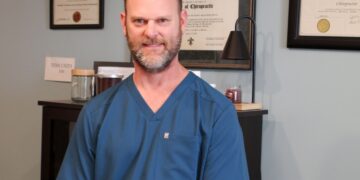 A professional portrait of Dr. Ronald Woods, a chiropractor with a short beard, wearing navy blue FIGS scrubs. He is standing in his office with his hands clasped in front of him. In the background, his diploma from Logan College of Chiropractic and his license from the State of Indiana are displayed on a light blue wall.