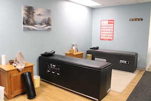 A serene chiropractic treatment room with light blue walls and wood-look flooring. Two black intersegmental traction tables, often called "roller tables," are positioned along the walls. Each table has a black bolster pillow at the head. A red-and-white eye chart hangs on the far wall, and a peaceful winter landscape painting is on the left wall. A small wooden side table holds a towel and supplementary therapy tools.