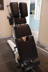 A close-up of a professional Hill DT decompression table or a specialized chiropractic drop table in a treatment room. The table features a white base with a series of black, high-density foam cushions designed to support the head, chest, and lower body. The table is shown in a partially tilted upright position, with a view into a hallway and a reception area in the background.