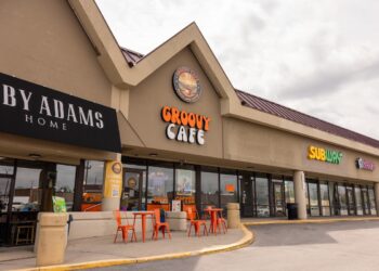 A daytime exterior shot of a one-story commercial strip mall under an overcast sky. The focal point is The Groovy Cafe, featuring a large, orange and white retro-style sign. Above it, a circular logo depicts a sun setting over a landscape with the words "PEACE LOVE COFFEE." Two small orange tables with matching chairs sit on the sidewalk in front of the glass storefront. To the left is a shop called "LIBBY ADAMS HOME" with a black awning, and to the right is a Subway restaurant.