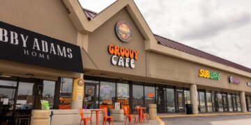 A daytime exterior shot of a one-story commercial strip mall under an overcast sky. The focal point is The Groovy Cafe, featuring a large, orange and white retro-style sign. Above it, a circular logo depicts a sun setting over a landscape with the words "PEACE LOVE COFFEE." Two small orange tables with matching chairs sit on the sidewalk in front of the glass storefront. To the left is a shop called "LIBBY ADAMS HOME" with a black awning, and to the right is a Subway restaurant.