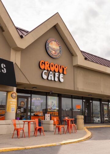 A daytime exterior shot of a one-story commercial strip mall under an overcast sky. The focal point is The Groovy Cafe, featuring a large, orange and white retro-style sign. Above it, a circular logo depicts a sun setting over a landscape with the words "PEACE LOVE COFFEE." Two small orange tables with matching chairs sit on the sidewalk in front of the glass storefront. To the left is a shop called "LIBBY ADAMS HOME" with a black awning, and to the right is a Subway restaurant.
