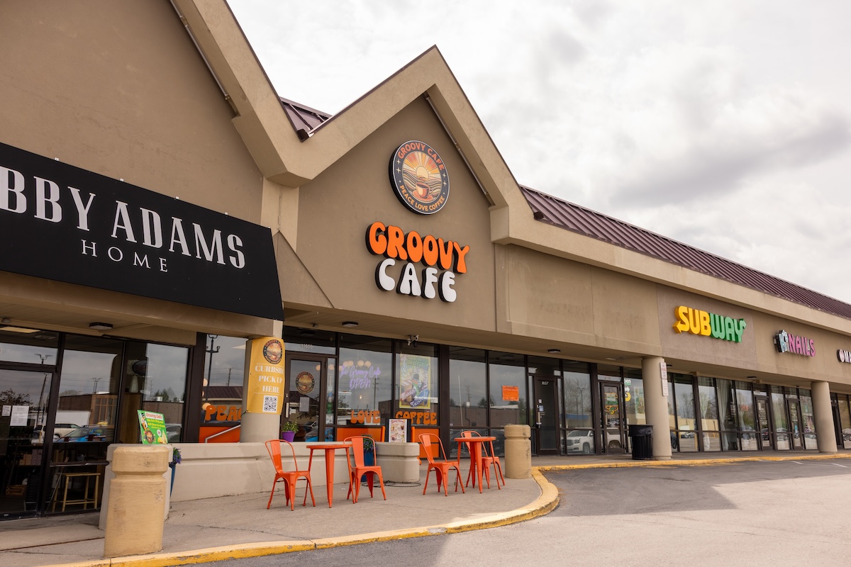 A daytime exterior shot of a one-story commercial strip mall under an overcast sky. The focal point is The Groovy Cafe, featuring a large, orange and white retro-style sign. Above it, a circular logo depicts a sun setting over a landscape with the words "PEACE LOVE COFFEE." Two small orange tables with matching chairs sit on the sidewalk in front of the glass storefront. To the left is a shop called "LIBBY ADAMS HOME" with a black awning, and to the right is a Subway restaurant.