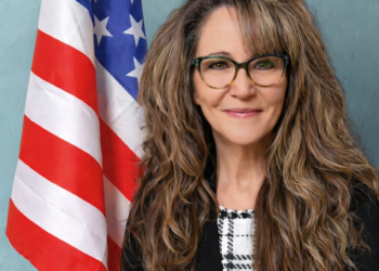 A woman with long brown and graying hair standing in front of the American flag.