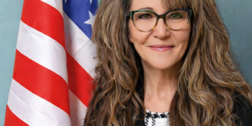 A woman with long brown and graying hair standing in front of the American flag.