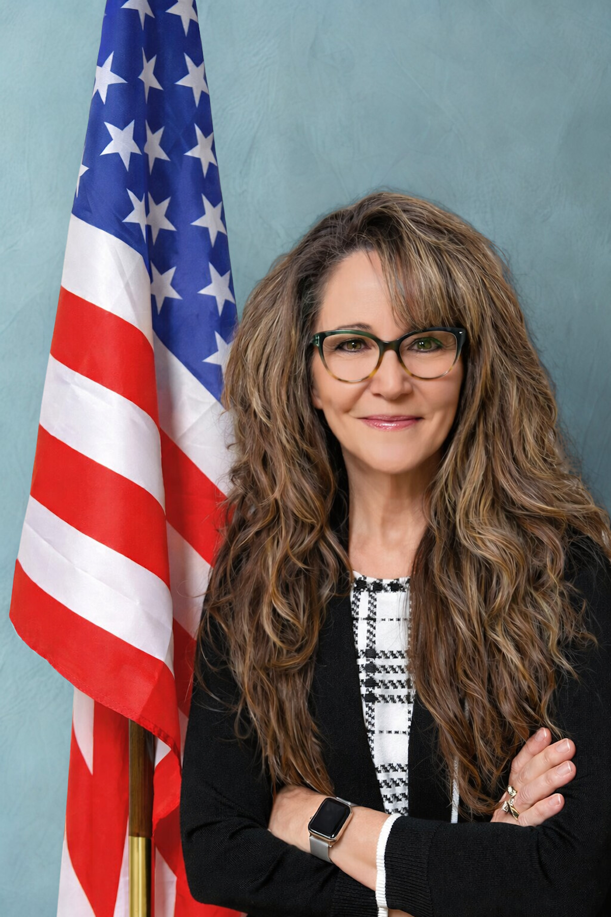 A woman with long brown and graying hair standing in front of the American flag.