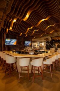 A medium full-shot of the upscale bar area at H Steakhouse in Westfield, Indiana. The focal point is a striking, undulating ceiling feature made of dark wood slats that ripple across the entire bar space, creating a sense of movement and warmth. The bar itself is a light-colored marble, arranged in a wide U-shape and surrounded by plush, cream-colored barrel-back stools with wooden legs. Two flat-screen TVs are mounted on a wall with matching dark wood vertical slats, displaying sports content. Behind the bar, shelves of premium spirits are visible under soft, warm lighting that accentuates the industrial-chic design of the space.