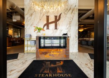 A wide-angle, eye-level shot looking through a dark doorway into the elegant entrance of H Steakhouse in Westfield, Indiana. The focal point is a large, stylized dark metal "H" logo mounted on a floor-to-ceiling white marble-patterned wall. Below the logo is a fireplace with a rustic, rusted-metal base. A crystal branch-style chandelier hangs from the ceiling, casting a warm glow. On the floor is a black welcome mat featuring the restaurant's logo and the word "STEAKHOUSE" in gold letters. To the left and right, blurred views of the modern dining areas are visible.