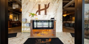 A wide-angle, eye-level shot looking through a dark doorway into the elegant entrance of H Steakhouse in Westfield, Indiana. The focal point is a large, stylized dark metal "H" logo mounted on a floor-to-ceiling white marble-patterned wall. Below the logo is a fireplace with a rustic, rusted-metal base. A crystal branch-style chandelier hangs from the ceiling, casting a warm glow. On the floor is a black welcome mat featuring the restaurant's logo and the word "STEAKHOUSE" in gold letters. To the left and right, blurred views of the modern dining areas are visible.