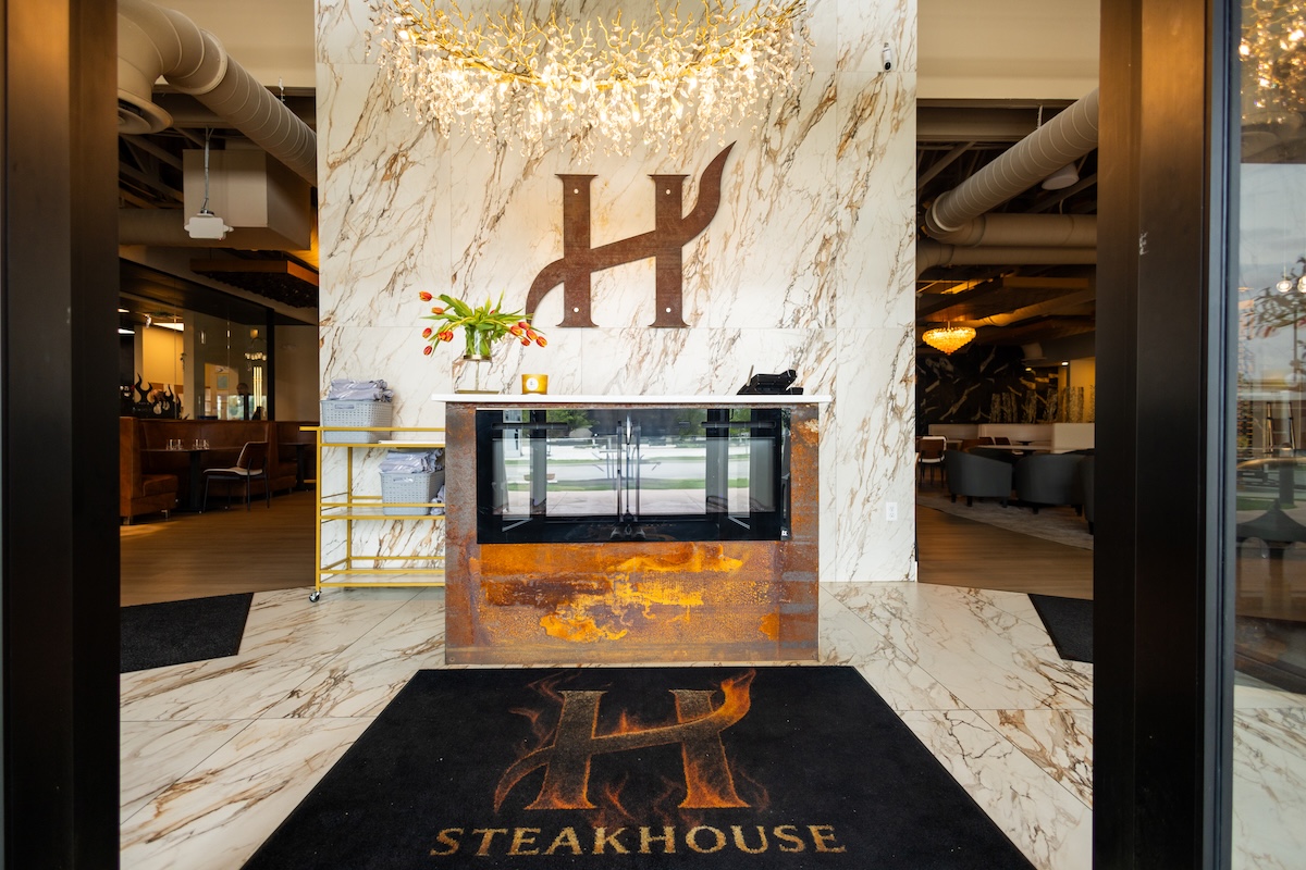 A wide-angle, eye-level shot looking through a dark doorway into the elegant entrance of H Steakhouse in Westfield, Indiana. The focal point is a large, stylized dark metal "H" logo mounted on a floor-to-ceiling white marble-patterned wall. Below the logo is a fireplace with a rustic, rusted-metal base. A crystal branch-style chandelier hangs from the ceiling, casting a warm glow. On the floor is a black welcome mat featuring the restaurant's logo and the word "STEAKHOUSE" in gold letters. To the left and right, blurred views of the modern dining areas are visible.