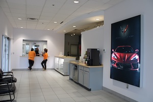 The interior of a car dealership featuring a customer service desk in the background. A large wall-mounted artwork of a red Chevrolet Corvette C8 is prominently displayed next to a coffee station.