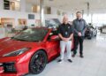 A professional portrait of two dealership employees standing by a new red Corvette C8 Z06. The showroom features high ceilings, glass walls, and other luxury vehicles in the background.