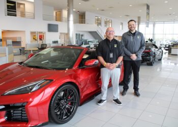 A professional portrait of two dealership employees standing by a new red Corvette C8 Z06. The showroom features high ceilings, glass walls, and other luxury vehicles in the background.