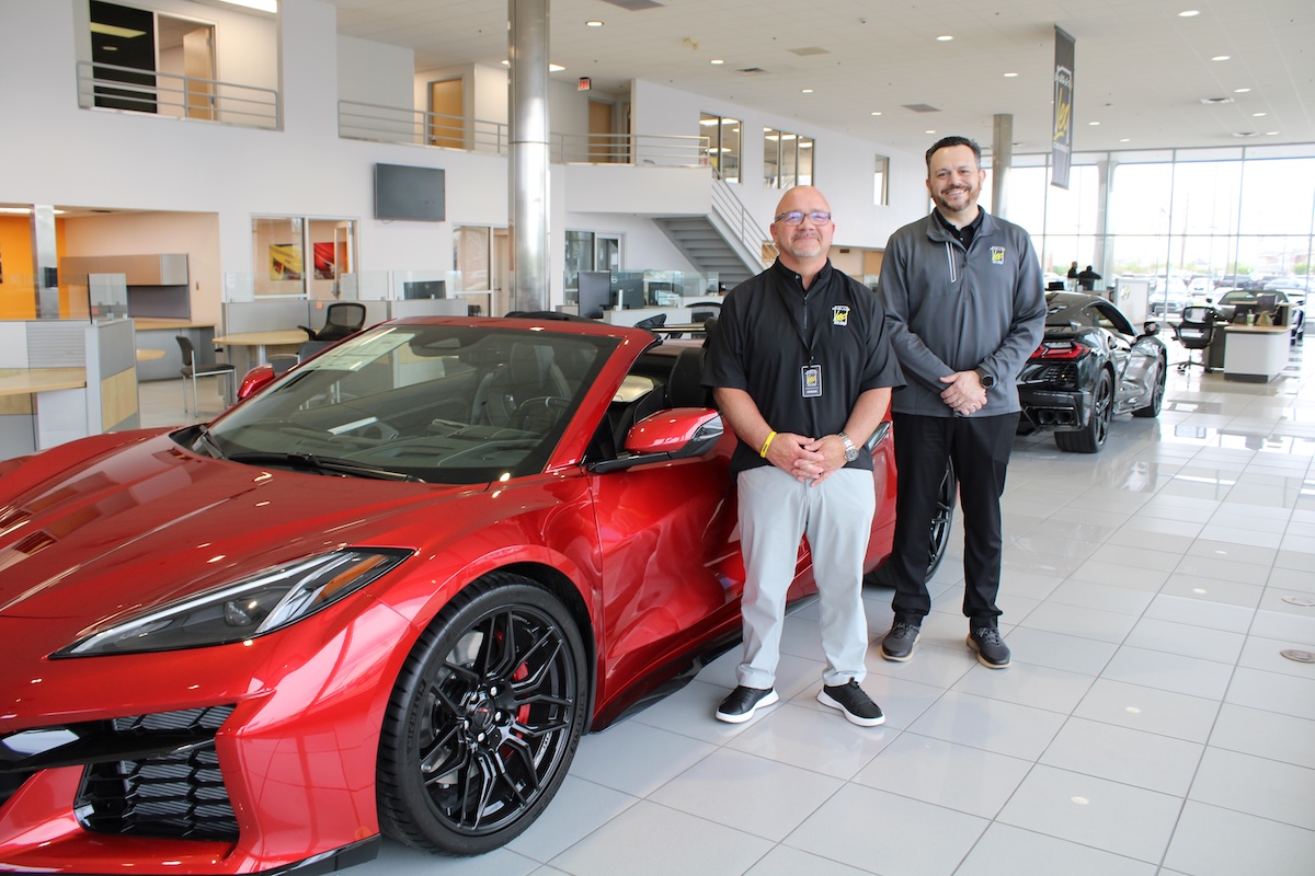 A professional portrait of two dealership employees standing by a new red Corvette C8 Z06. The showroom features high ceilings, glass walls, and other luxury vehicles in the background.