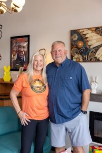 A smiling man and woman stand close together in a warmly lit cafe interior. The woman, with long blonde hair, wears an orange t-shirt featuring the circular "PEACE LOVE COFFEE" logo of The Groovy Cafe. The man, with short grey hair, wears a blue button-down short-sleeved shirt and grey shorts. Behind them, eclectic decor includes a framed portrait of Martin Luther King Jr., a large piece of butterfly wing art, a yellow Peeps-shaped decoration, and a small electric fireplace.