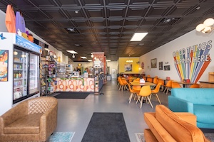 The interior of a brightly lit cafe with a high, black-tiled ceiling and a long, narrow layout. On the right wall, a colorful mural features an explosion of vibrant, rainbow-colored lines emanating from the corner with the words "GOOD THINGS HAPPEN HERE" written in stylized letters. The seating includes several small square tables paired with bright orange and yellow chairs, as well as a plush blue sofa and an orange armchair in the foreground. A service counter with a patterned facade and various coffee-making equipment is visible in the background, along with shelves of merchandise and a refrigerator stocked with drinks.