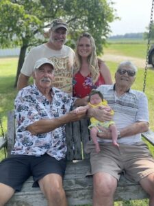 A multi-generational family photo taken outdoors on a sunny day. Two older men are seated on a rustic wooden porch swing; the man on the right holds a newborn baby wearing a yellow outfit. Standing behind the swing are a younger man in a baseball cap and graphic tee and a woman with long blonde hair in a red floral dress. Everyone is smiling, and the background shows a lush green yard and trees.