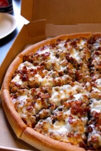 A close-up, high-angle shot of a Godfather's Pizza "All-Meat Combo" pizza inside its open cardboard delivery box. The pizza features a thick, golden-brown "Original" crust and is heavily loaded with various meat toppings including pepperoni, ham, beef, sausage, Italian sausage, and bacon, all smothered in melted mozzarella cheese. A white plate is partially visible in the blurred background to the left.
