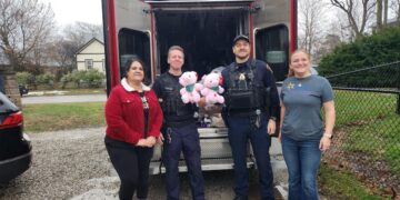 Two police officers in uniform and two women stand behind an open black IMPD patrol vehicle filled with donated toys. One officer holds two pink teddy bears with green bows. The woman on the left wears a red corduroy jacket with a white collar, and the woman on the right wears a grey t-shirt. The group is smiling on a paved driveway in a residential area.