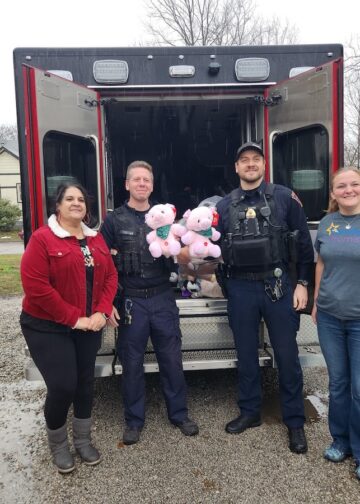 Two police officers in uniform and two women stand behind an open black IMPD patrol vehicle filled with donated toys. One officer holds two pink teddy bears with green bows. The woman on the left wears a red corduroy jacket with a white collar, and the woman on the right wears a grey t-shirt. The group is smiling on a paved driveway in a residential area.