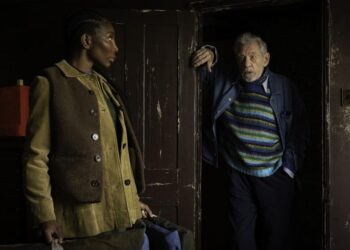 A medium shot in a dimly lit, rustic room features a young Black woman, Lori (Michaela Coel), standing in profile on the left. She wears a brown vest over a tan corduroy shirt and looks toward an older white man, Julian (Ian McKellen). Julian leans against a dark wooden doorframe on the right, wearing a vibrant blue, green, and yellow striped sweater under a blue jacket. The weathered walls and dark tones suggest a somber yet intimate setting.
