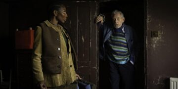 A medium shot in a dimly lit, rustic room features a young Black woman, Lori (Michaela Coel), standing in profile on the left. She wears a brown vest over a tan corduroy shirt and looks toward an older white man, Julian (Ian McKellen). Julian leans against a dark wooden doorframe on the right, wearing a vibrant blue, green, and yellow striped sweater under a blue jacket. The weathered walls and dark tones suggest a somber yet intimate setting.