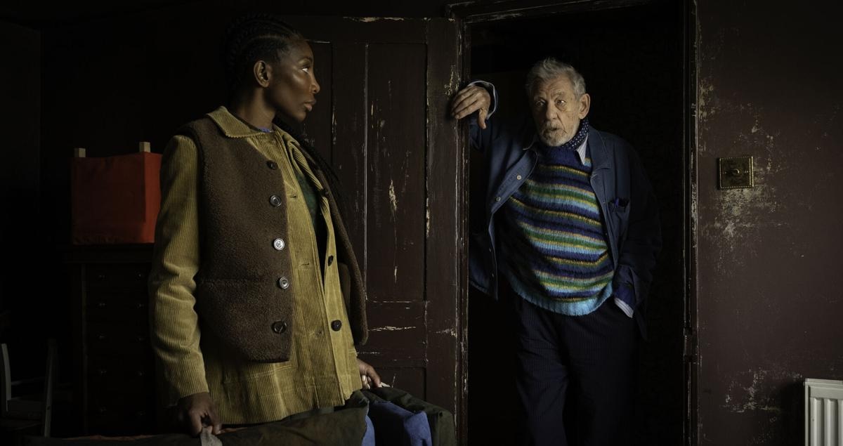 A medium shot in a dimly lit, rustic room features a young Black woman, Lori (Michaela Coel), standing in profile on the left. She wears a brown vest over a tan corduroy shirt and looks toward an older white man, Julian (Ian McKellen). Julian leans against a dark wooden doorframe on the right, wearing a vibrant blue, green, and yellow striped sweater under a blue jacket. The weathered walls and dark tones suggest a somber yet intimate setting.