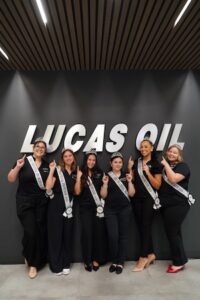 A medium full-shot of six young women, serving as 2026 500 Festival Princesses, posing together in front of a large, illuminated "LUCAS OIL" logo on a dark grey wall. The women are all dressed in matching black outfits and each wears a silver tiara and a white sash that reads "500 FESTIVAL PRINCESS 2026." They are all smiling and holding their right index fingers up in a "#1" gesture. The setting is modern and professional, likely the new Lucas Oil Global Headquarters in Indianapolis.