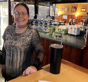A woman with glasses and dark hair pulled back stands smiling behind a bar. She is wearing a grey and black patterned t-shirt over a black long-sleeved mesh top. On the bar counter in front of her is a tall black tumbler with a straw. Behind her, rows of inverted blue-rimmed margarita glasses are neatly arranged on the bar, and wood-paneled walls with small framed pictures are visible in the background.