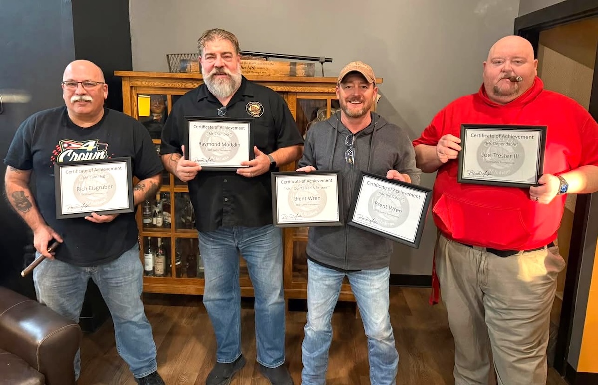 Four men stand in a row in a room with a warm, casual atmosphere, each holding framed "Certificate of Achievement" awards. From left to right, the men are Rich Eisgruber, Raymond Modglin, Brent Wren (holding two certificates), and Joe Trester III. Behind them is a wooden cabinet stocked with various bottles, and the overall setting suggests a social club or lounge environment.