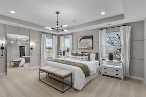 A wide-angle shot of a luxurious master bedroom in an M/I Homes model. The room features grey paneled walls and a tray ceiling with a modern black sputnik-style chandelier. A king-sized bed with crisp white linens and decorative pillows is flanked by two white three-drawer nightstands topped with black lamps. To the left, a large floor-to-ceiling mirror leans against the wall. A wooden bench with a white cushion sits at the foot of the bed on a light-colored, patterned carpet. Natural light streams in through three large windows dressed with white curtains.