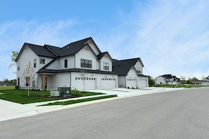 A low-angle, wide-perspective shot of a newly constructed modern farmhouse-style townhome building by M/I Homes in the Bonterra community. The two-story building features white vertical board-and-batten siding, black gabled roofs, and black-framed windows. Three separate units are visible, each with a white two-car garage and a small concrete driveway. A manicured green lawn with small, young trees and a grey paved street are in the foreground, all set against a bright, pale blue sky.