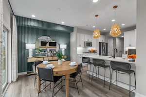 A wide-angle interior shot of a modern, open-concept kitchen and dining space in an M/I Homes townhome. In the foreground, a round light-wood dining table with black chairs is set for four. Behind it, a striking accent wall features dark teal vertical fluted paneling, a large round mirror, and a wood console table with white lamps. The kitchen in the background features white cabinetry, a white quartz island with three black mesh barstools, and two large, woven wood pendant lights. Light-colored wood-look flooring runs throughout the space.