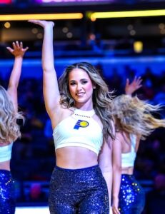 An Indiana Pacers dancer with blonde highlighted hair performs mid-routine on the basketball court. She is wearing a white high-neck crop top with the Pacers logo and shimmering navy blue sequined leggings. Her right arm is raised straight up, while other dancers in the same uniform are visible in the background, their movement creating a slight blur against the arena lights.