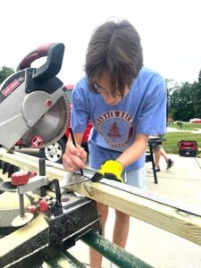 A young person with brown hair, wearing yellow work gloves and a light blue t-shirt with a tree logo, focuses on marking a piece of lumber with a pencil. They are working at a sliding miter saw outdoors on a sunny day. Wood shavings are visible on the saw’s base, and another person can be seen working in the background of the paved area.