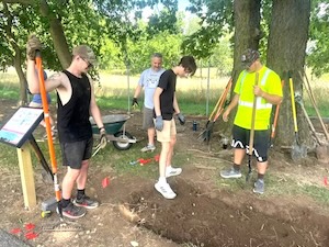 Three young men and an older man are working on an outdoor landscaping project on a sunny day. In the foreground, a youth in a black tank top holds a shovel near a freshly dug trench, while another in a black t-shirt and tan shorts stands in the dirt. On the right, a man in a high-visibility yellow polo shirt and a camouflage hat holds a long-handled tool. A green wheelbarrow and several other shovels leaning against a large tree are visible in the background.
