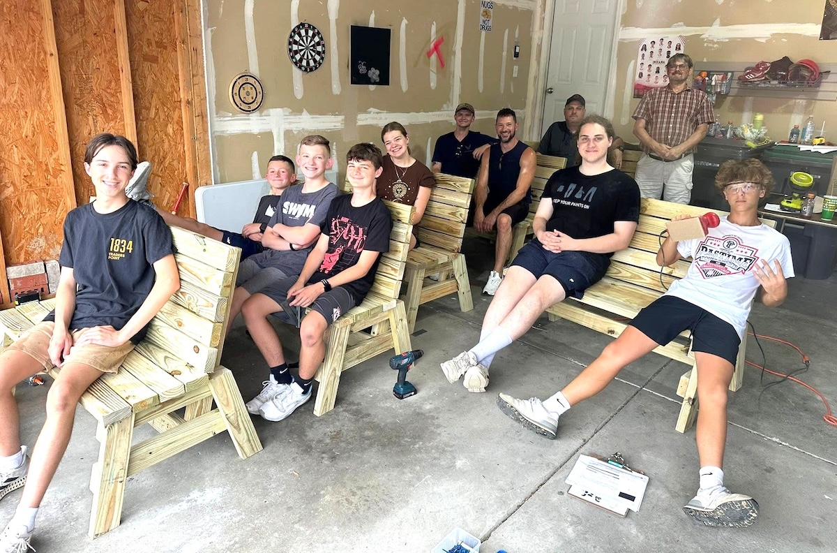 In a spacious garage workshop, a group of seven young people and three men pose for a photo. They are seated on four sturdy, handmade wooden benches with slatted backs and seats. The garage features exposed wall studs, a dartboard, and a whiteboard. One youth in the center wears a black shirt with a red graphic, and another on the far right holds a red object, while a man in a plaid shirt stands in the background near a workbench with tools.