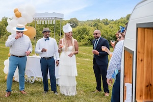 A group of five well-dressed people are gathered outdoors in a grassy field, celebrating near a white mobile bar cart. A woman in a white tiered dress and a matching fascinator stands in the center, smiling. To her left, a man in a white shirt and blue pants takes a drink, while another man in a bow tie and fedora stands beside her. To her right, a man in a dark suit and sunglasses looks on. The background features lush green trees under a bright sky, with a cluster of white and gold balloons adding to the festive atmosphere.